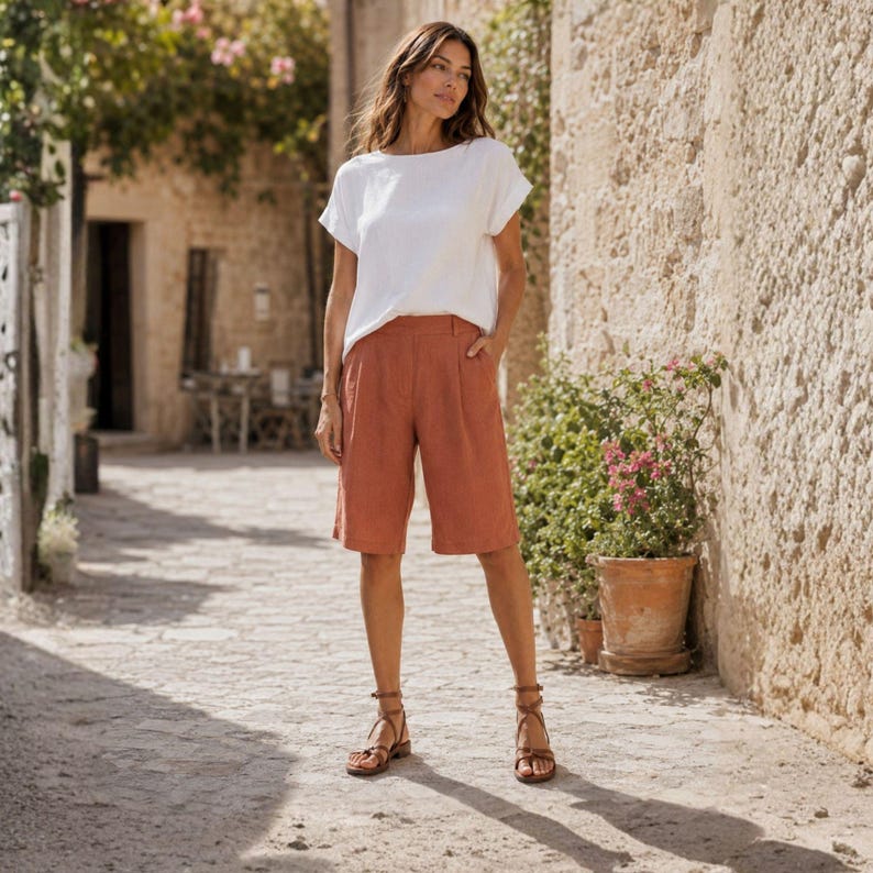 May include: A white short-sleeved top with a round neckline and loose fit paired with knee-length terracotta-colored shorts featuring a button closure and side pockets, worn with brown strappy sandals with ankle straps and a buckle closure.