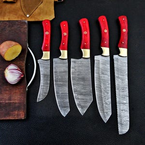 May include: A collection of six kitchen knives featuring red handles and patterned blades. The handles have brass bolsters and are riveted. The blades display a distinctive pattern. A wooden chopping board, a potato, and an onion are in the background.