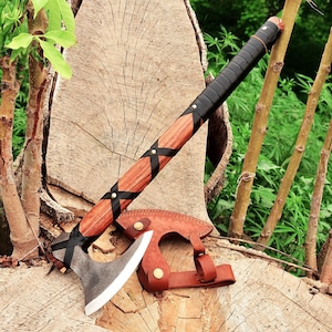 Peut inclure: Une hache artisanale avec un manche en bois enveloppé de cuir noir et orné de lanières noires. La tête de la hache est argentée avec une lame tranchante. Un étui en cuir marron protège la lame. La hache repose sur une souche d'arbre.