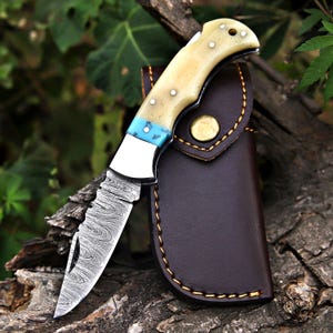 May include: A folding knife with a patterned blade and a cream-coloured handle with blue accents. The knife is paired with a brown leather sheath. The blade has a unique, textured design. The knife is resting on a piece of wood.