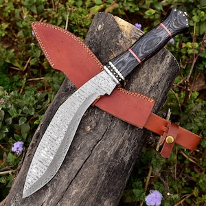 May include: A handcrafted kukri knife with a curved, patterned steel blade and a dark wooden handle. The handle is accented with red stripes and brass studs. It rests in a brown leather sheath with a belt loop, displayed on a wooden log.