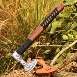 May include: A handcrafted axe with a wooden handle and a black leather-wrapped grip. The axe head features intricate white designs on a silver-coloured blade. A brown leather sheath protects the blade. The axe is displayed on a tree branch.