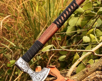 May include: A handcrafted axe with a wooden handle and a black leather-wrapped grip. The axe head features intricate white designs on a silver-colored blade. A brown leather sheath protects the blade. The axe is displayed on a tree branch.