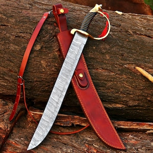 May include: A large, curved sword with a patterned blade is in a red leather sheath. The sword has a gold-coloured hilt and a black handle. A red leather strap is attached to the sheath. The sword is displayed on a textured, brown wooden surface.