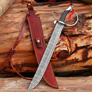 May include: A Damascus steel sword with a leather-wrapped handle and a silver guard. The blade is sheathed in a brown leather scabbard with a shoulder strap. The sword and scabbard are displayed on a wooden surface.