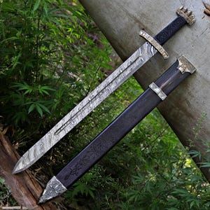 May include: A Viking sword with a patterned blade and a black handle, shown with its matching black sheath. Both the sword and sheath have detailed metalwork and designs. The sword is displayed on a tree trunk, surrounded by greenery.