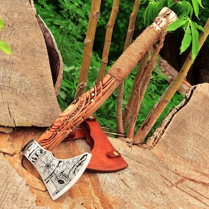 May include: A handcrafted axe with a wooden handle wrapped in twine. The axe head is silver with intricate etched designs, including a compass symbol. A brown leather sheath protects the blade. The axe rests on a wooden surface with green foliage in the background.