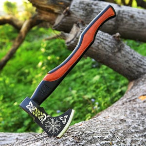 May include: A Viking axe with a black and orange handle. The axe head has gold and white Celtic knot designs and a white symbol. The axe is displayed on a tree branch.