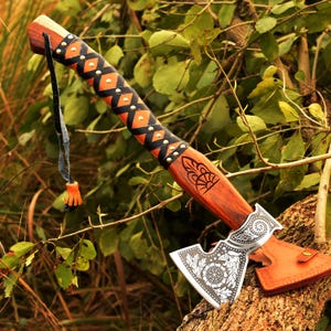 May include: A decorative axe with a wooden handle and a detailed blade. The handle is wrapped in black and orange leather with gold rivets. The blade features an etched floral design and comes with a leather sheath.