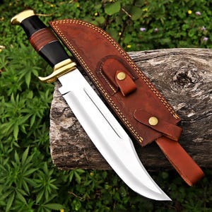 May include: A large, polished steel knife with a curved blade, brass accents, and a black and brown handle. It rests beside a brown leather sheath with decorative stitching and brass closures. The knife is designed for outdoor use.