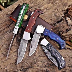 May include: A collection of folding knives with diverse handle designs and blade patterns. The handles are made of wood, green, and blue materials. The blades have a textured pattern. A brown leather case is also visible.