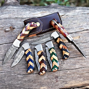 May include: A collection of folding knives with patterned wooden handles in blue, brown, and green. The blades have a textured design. A brown leather sheath is also visible.