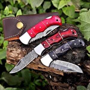 May include: Three folding knives with wooden handles in red, dark red, and black, displayed on a wooden surface. The blades have a unique pattern. A brown leather case with a gold button is also present.