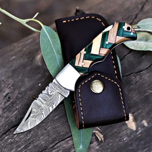 May include: A folding knife with a patterned blade and a handle made of green, tan, and light wood pieces. The knife is in a black leather sheath with a brass snap closure. The blade has a unique, swirling pattern.