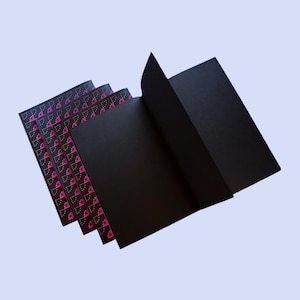 Pode incluir: Cadernos pretos com um padrão floral rosa nas bordas. Um dos cadernos está aberto, revelando páginas pretas em branco. Os cadernos estão dispostos sobre um fundo azul claro.