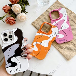 Könnte beinhalten: Drei Handyhüllen mit abstrakten Designs. Eine Hülle ist weiß mit schwarzen und grauen Wirbeln und dem Text "Mrs. H". Eine andere ist orange mit weißen Wirbeln und dem Text "dan". Die dritte ist rosa mit weißen Wirbeln und dem Text "Madu".