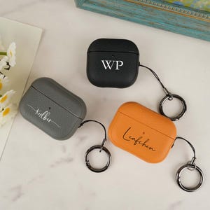 May include: Three personalised AirPod cases in black, grey, and orange leather. The black case has "WP" in white, the grey case has "Kilbir" in white, and the orange case has "Leafchen" in white. Each case has a silver-coloured keychain.