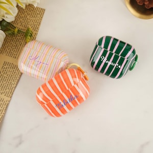 Könnte beinhalten: Drei AirPod-Hüllen mit farbenfrohen Streifendesigns. Eine Hülle ist orange und weiß, eine andere grün und rosa, und die dritte ist pastellfarben. Auf jeder Hülle steht ein Name: "Tabitha", "Megan" und "Megan".