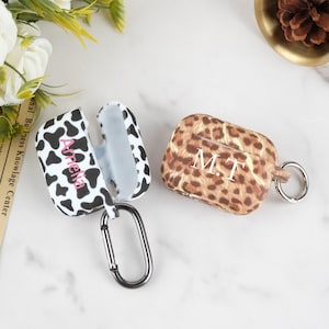 Könnte beinhalten: Zwei personalisierte AirPod-Hüllen. Eine Hülle ist weiß mit schwarzen Kuhflecken und dem Namen "Amelia" in Rosa. Die andere Hülle hat einen braunen Leopardenmusterdruck mit den Initialen "M.T." in Weiß. Beide Hüllen haben einen silbernen Karabinerhaken.