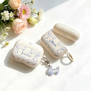 Könnte beinhalten: Cremefarbene AirPod-Hüllen mit blauen Dackel-Illustrationen und den Namen "Jordan" und "Madu". Eine Hülle hat einen Schlüsselanhänger mit einem passenden Hundeanhänger. Eine passende AirPod-Hülle ist ebenfalls sichtbar. Die Hüllen befinden sich auf einer weißen Oberfläche mit Blumen.