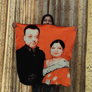 May include: An orange knitted blanket featuring a portrait of two people. The portrait is rendered in black, white, and shades of orange, with detailed facial features and clothing. The blanket is held up by a person.