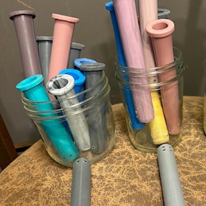 Puede incluir: Un surtido de objetos cilíndricos de colores, probablemente tapones de botella, en varios tonos de rosa, azul, gris y amarillo. Se exhiben en frascos de vidrio transparente, con algunos de pie solos. Los tapones tienen una parte superior redondeada y una base acampanada.