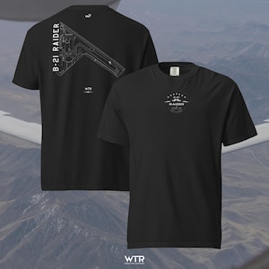 May include: A black t-shirt featuring a white graphic print. The back displays a technical line drawing of the B-21 Raider aircraft with the text B-21 RAIDER. The front chest area features a smaller circular logo with the text B-21 RAIDER.