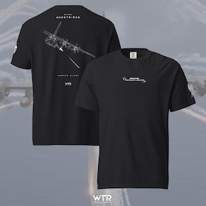 May include: A black short-sleeved t-shirt featuring a white line art graphic of an AC-130J Ghostrider aircraft on the back with the text "AC-130J GHOSTRIDER" and "DANGER CLOSE". The front chest displays "BOEING LOCKHEED MARTIN" text in white.