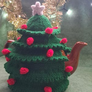 May include: A green knitted tea cosy shaped like a Christmas tree, adorned with red pom-poms and a white star. The cosy covers a brown teapot, perfect for festive tea times.