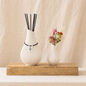 Könnte beinhalten: Zwei weiße, gerippte Keramikvasen auf einem Holzblock. Eine Vase enthält schwarze Schilfrohre, die andere Trockenblumen. Eine schwarze Kordel mit einem kleinen Anhänger hängt an der größeren Vase. Der Hintergrund ist ein neutralfarbener Stoff.