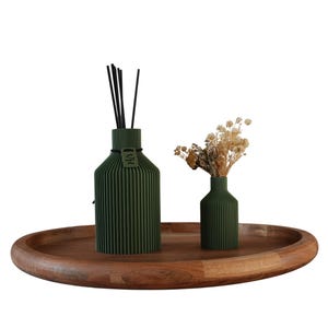 Könnte beinhalten: Ein dunkelgrüner Schilf-Diffusor mit schwarzen Schilfstäben und eine kleine grüne Vase mit getrockneten Blumen stehen auf einem ovalen Holztablett. Der Diffusor und die Vase haben ein geripptes Design.