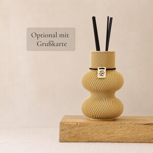 Könnte beinhalten: Ein beigefarbener Raumduft-Diffusor mit einem texturierten, gerippten, sanduhrförmigen Körper und vier schwarzen Stäbchen. Ein kleines rechteckiges Etikett mit den Buchstaben SL ist mit einem schwarzen Gummiband am Hals befestigt. Der Text Optional mit Grußkarte ist sichtbar.