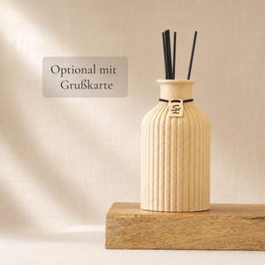 Könnte beinhalten: Eine beige, geriffelte Keramikflasche für einen Raumduft mit engem Hals und einem kleinen Holzanhänger mit den Buchstaben SL. Fünf dünne schwarze Stäbchen stecken oben im Flakon. Ein rechteckiges Schild links zeigt den Text Optional mit Grußkarte.