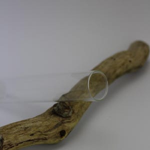 Könnte beinhalten: Ein klares Glasrohr liegt auf einem Stück Treibholz. Das Treibholz ist hellbraun und hat eine strukturierte Oberfläche. Das Glasrohr ist zylindrisch und hat ein glattes, transparentes Aussehen. Der Hintergrund ist einfarbig weiß.