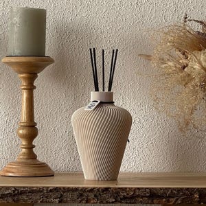 Könnte beinhalten: Ein beigefarbener Raumduft-Diffusor mit spiralförmiger, gerippter Textur und vier schwarzen Stäbchen, die oben eingesteckt sind. Ein kleines weißes Etikett mit schwarzem Text ist am Flaschenhals befestigt. Der Diffusor steht neben einem Kerzenhalter.