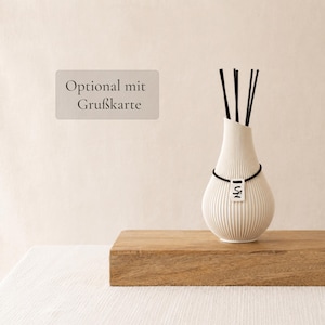 Könnte beinhalten: Ein weißer, gerippter Diffuser mit rundem Boden und engem Hals. Ein schwarzes Gummiband umschließt den Hals und hält ein kleines, weißes, rechteckiges Schild mit schwarzer Aufschrift. Fünf schwarze Stäbchen ragen aus der Öffnung.