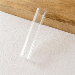 Könnte beinhalten: Ein klarer, zylindrischer Glasstab, an beiden Enden offen. Der Stab ist gerade mit einem gleichmäßigen Durchmesser. Der Hintergrund umfasst eine Holzoberfläche und weißen Stoff. Geeignet für Dekoration oder Experimente.