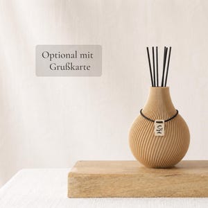 Könnte beinhalten: Ein beigefarbener Raumduft-Diffusor mit gerippter, kugelförmiger Basis und schwarzen Stäbchen. Eine schwarze Kordel umschließt den Hals und hält ein kleines rechteckiges Schild mit den Buchstaben SL. Der Text Optional mit Grußkarte ist zu sehen.
