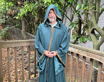 May include: A person wearing a long, teal hooded robe with gold trim and a dark green lining. The robe has long sleeves and a sash tied at the waist. The person is standing on a wooden deck with leaves scattered around.