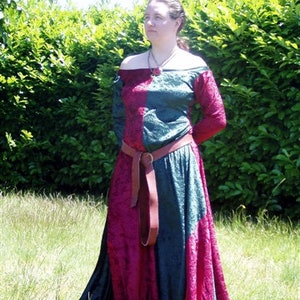 May include: A medieval-style dress with a burgundy and teal color block design. The off-the-shoulder dress has long sleeves and a wide brown belt. The dress is worn outdoors in front of a green hedge.