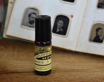 May include: A small black roller bottle with a gold label that reads "Apothecary Scents Smoke & Mirrors" with the words "Organic rice bran oil" and "External use only" on the label. The bottle is sitting on a wooden surface in front of an old photo album.