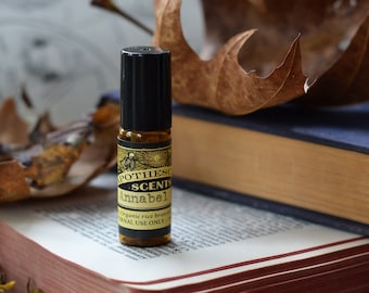 Pode incluir: Um pequeno frasco de vidro com uma tampa preta e um rótulo dourado que diz "Pothesies Scents Annabel" e "Óleo de farelo de arroz orgânico para uso externo somente". O frasco está sentado em uma pilha de livros com uma capa de couro marrom e uma capa de tecido azul. Há uma folha marrom e um cachimbo de madeira no fundo.