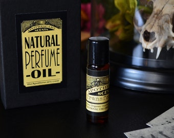 May include: A small, dark brown bottle with a gold label that reads "Apothecary Scents - Natural Perfume Oil - theapothecary.com". The bottle is sitting on a black surface next to a black box with a gold label that reads "Apothecary Scents - Natural Perfume Oil - theapothecary.com".