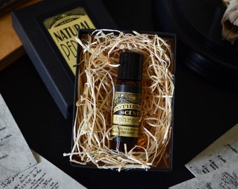 Pode incluir: Um pequeno frasco de vidro com um rótulo preto e letras douradas. O rótulo diz "POTHE SCENT Aphrodite" e "TAKE ONE DROP ONLY". O frasco está em uma caixa cheia de papel picado.