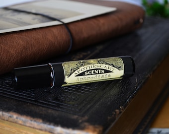Pode incluir: Um frasco de roll-on preto e dourado com um rótulo que diz "Apothescary Scents Aphrodisia". O frasco está em cima de um livro encadernado em couro castanho.