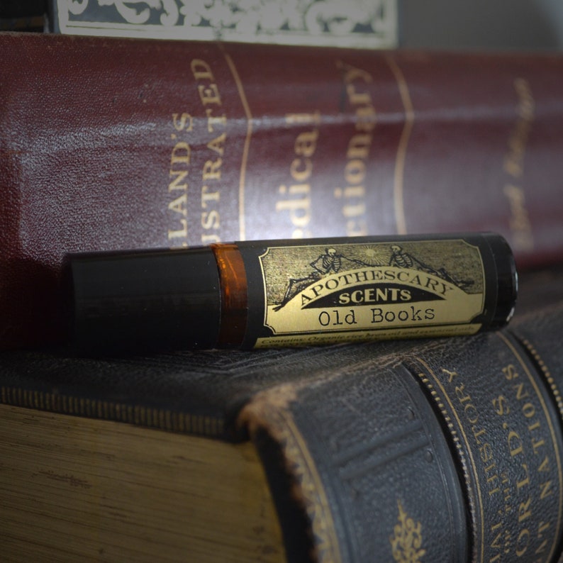Puede incluir: Una botella de vidrio marr&oacute;n con una etiqueta dorada que dice "Apothecary Scents Old Books". La botella est&aacute; encima de una pila de libros antiguos.