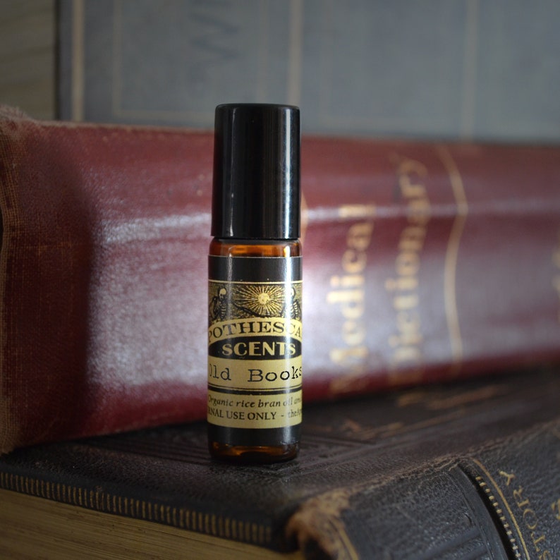 Puede incluir: Un frasco de vidrio marr&oacute;n con forma de rodillo con una tapa negra y una etiqueta que dice "Apothecary Scents Old Books" con el texto "organic rice bran oil" y "external use only". El frasco est&aacute; sobre una pila de libros antiguos.