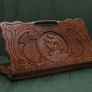 May include: A wooden backgammon set with a carved ship design. The dark brown wooden case features a handle and intricate floral and geometric patterns. The central design is a detailed relief of a sailing ship.