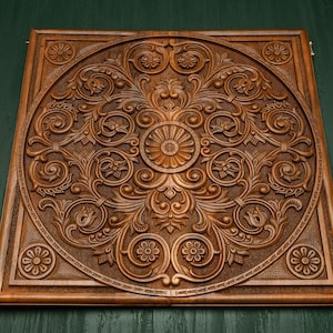 May include: A square, wooden backgammon board with intricate, carved floral and scrollwork designs. The board is a warm brown color and features a central circular motif surrounded by detailed patterns. The corners have small, decorative floral elements.
