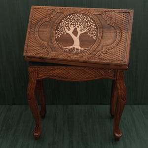 May include: A wooden backgammon set with a carved tree of life design on the lid. The set is brown with a detailed lattice pattern and rests on a matching wooden stand with curved legs. The set is a classic game.